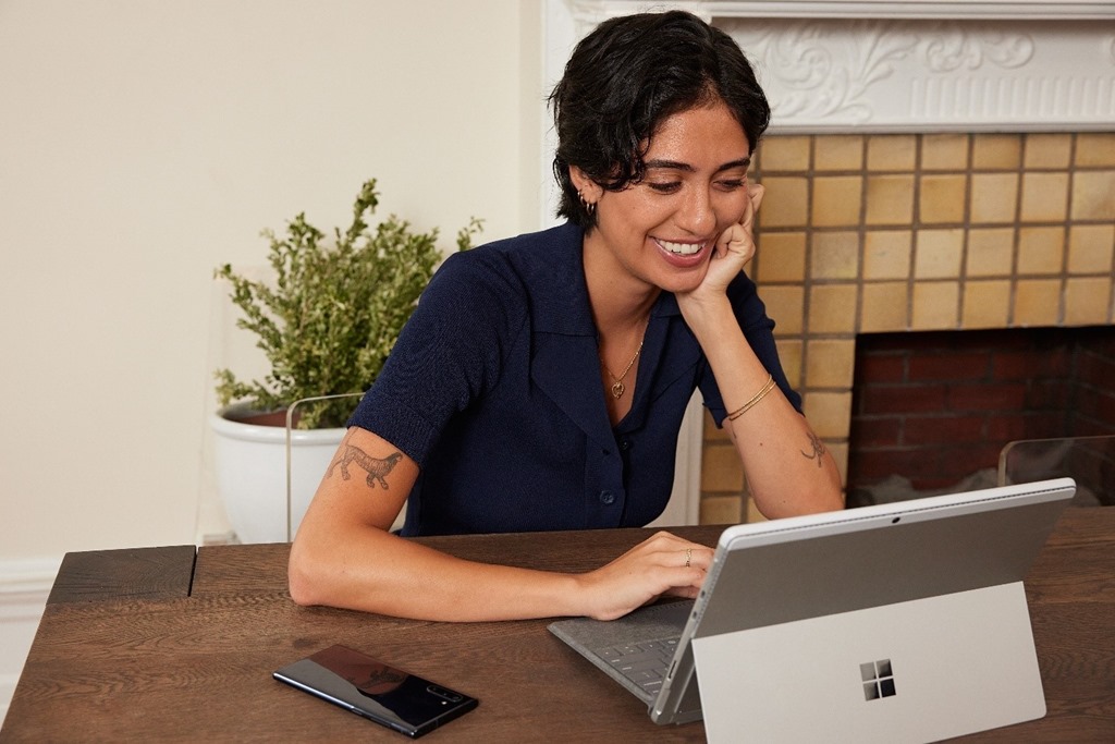 Жінка сидить за столом з планшетом Microsoft та мобільним телефоном. Вона посміхається на планшеті.
