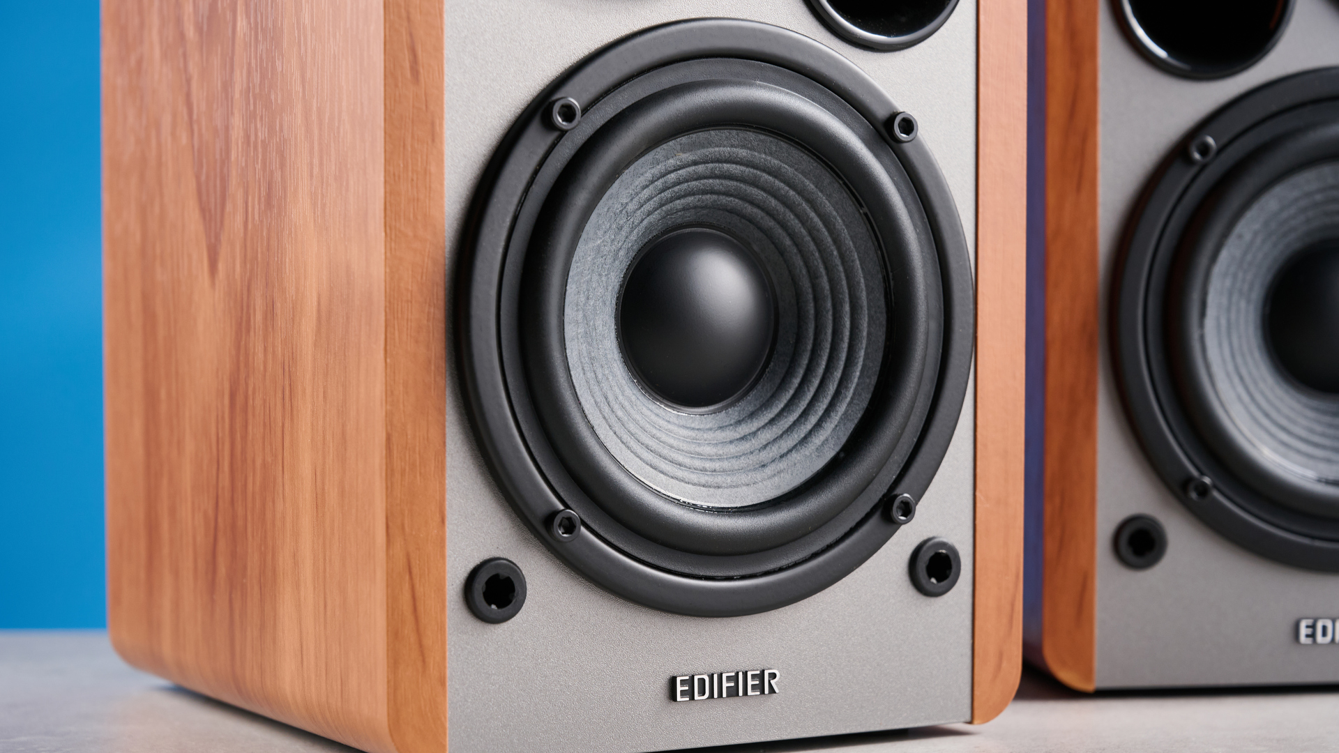 Дивнори для книжкових полиць Edifier R1280T у деревному коричневому кольорі, сфотографованому на синьому тлі, показуючи циферблат регулювання гучності, внутрішні драйвери та твітери, входи RCA та AUX та віддалені