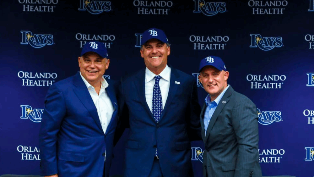 Троє членів нової групи власників Tampa Bay Rays позують разом у капелюхах команди на тлі прес-конференції з логотипами Rays та Orlando Health.